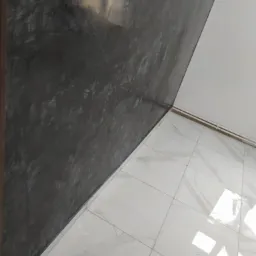 Ściana z efektem betonu w odcieniach szarości, kontrastująca z białymi płytkami podłogowymi imitującymi marmur i białym sufitem, widoczne rury grzewcze przy suficie.