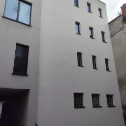 Jasna elewacja budynku z rzędami okien, widok z dołu. Obok budynek z wejściem i oknami z żaluzjami. W tle widać fragment sąsiedniego budynku.