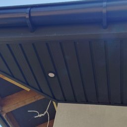 SbudDom - Ciemna podbitka dachowa z oświetleniem punktowym, widok z dołu, fragment konstrukcji drewnianej i orynnowanie. Detal wykończenia.