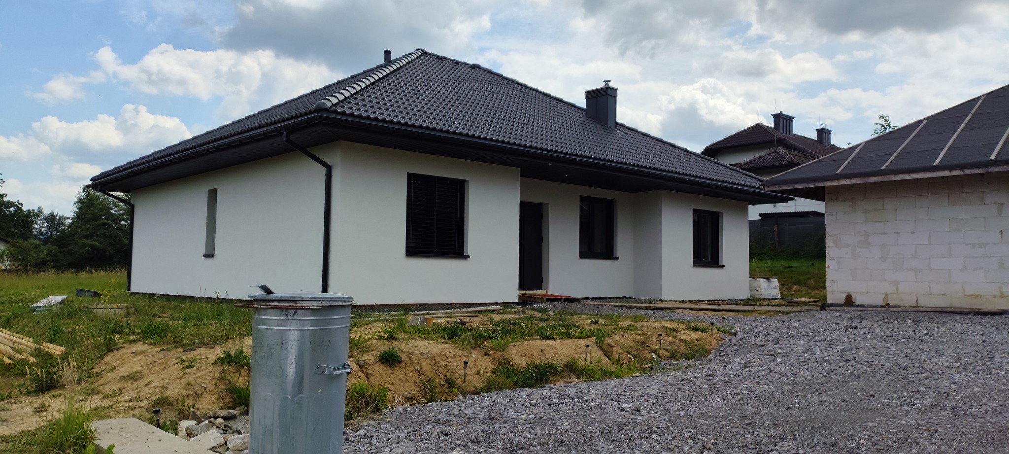 Nowoczesny dom jednorodzinny z ciemną dachówką i jasną elewacją, widok z zewnątrz, teren wokół domu w trakcie zagospodarowania, częściowo wykończony budynek gospodarczy.