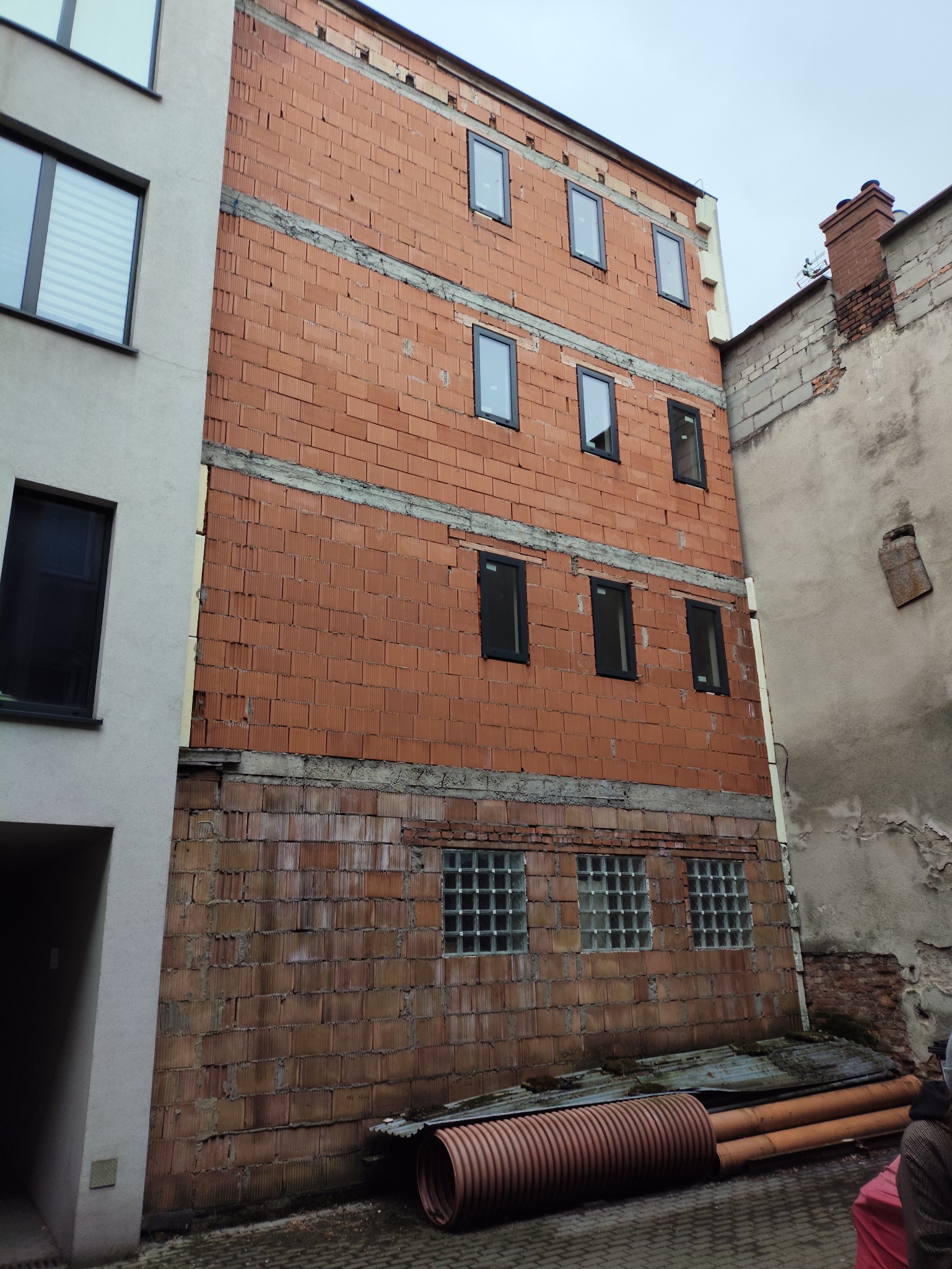 Budynek w trakcie budowy z czerwonej cegły, widoczne okna i rury na dole, sąsiedni budynek z oknami.