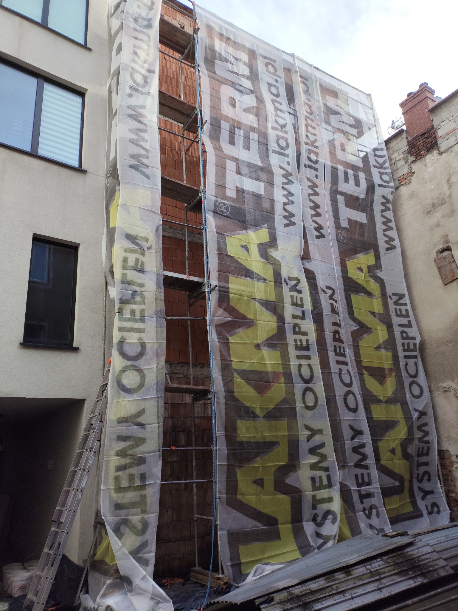 Budynek w trakcie prac elewacyjnych, osłonięty siatką z reklamą, widoczne rusztowania i drabina. Ceglana ściana w trakcie ocieplania.