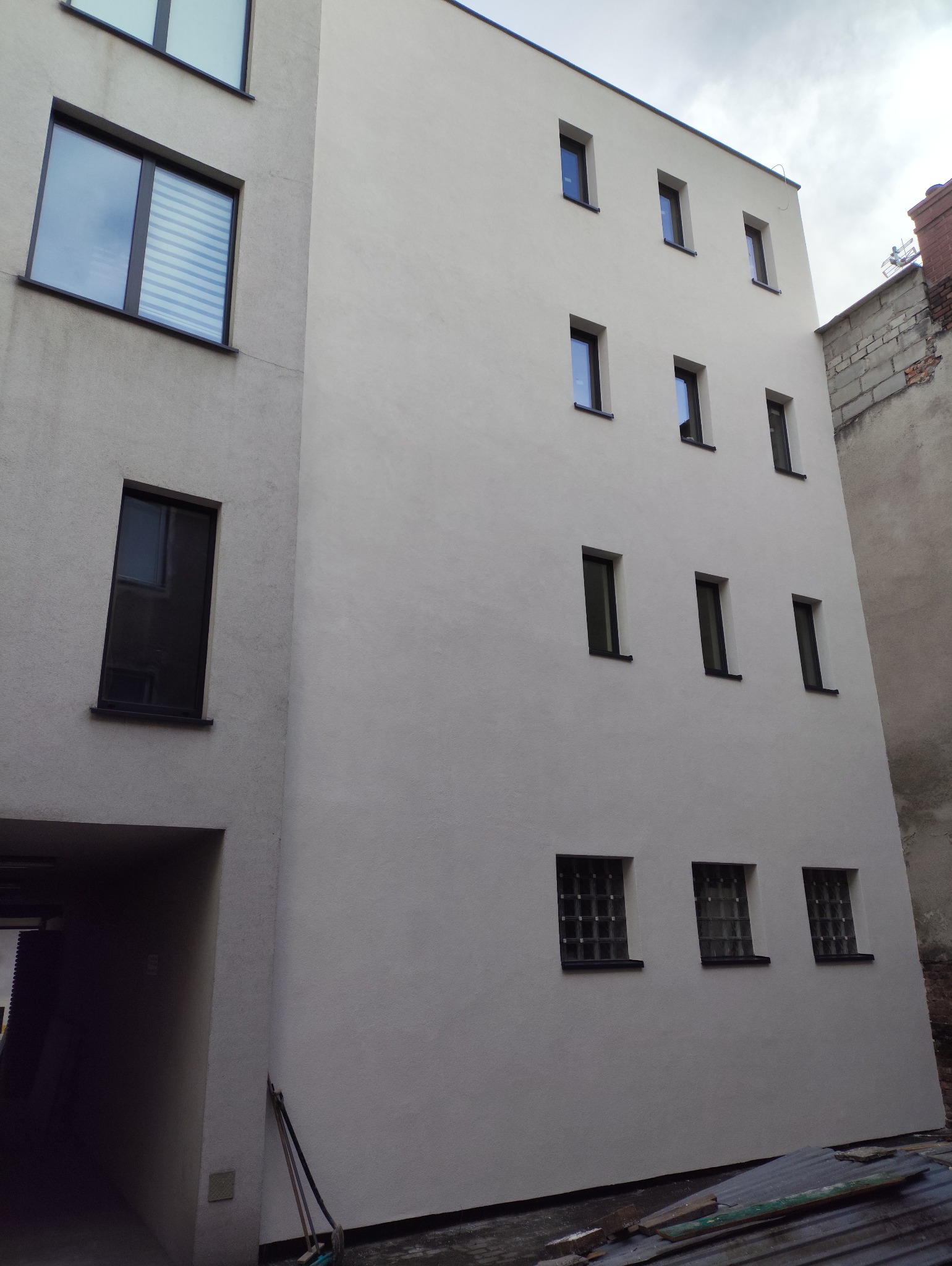 Jasna elewacja budynku z rzędami okien, widok z dołu. Obok budynek z wejściem i oknami z żaluzjami. W tle widać fragment sąsiedniego budynku.