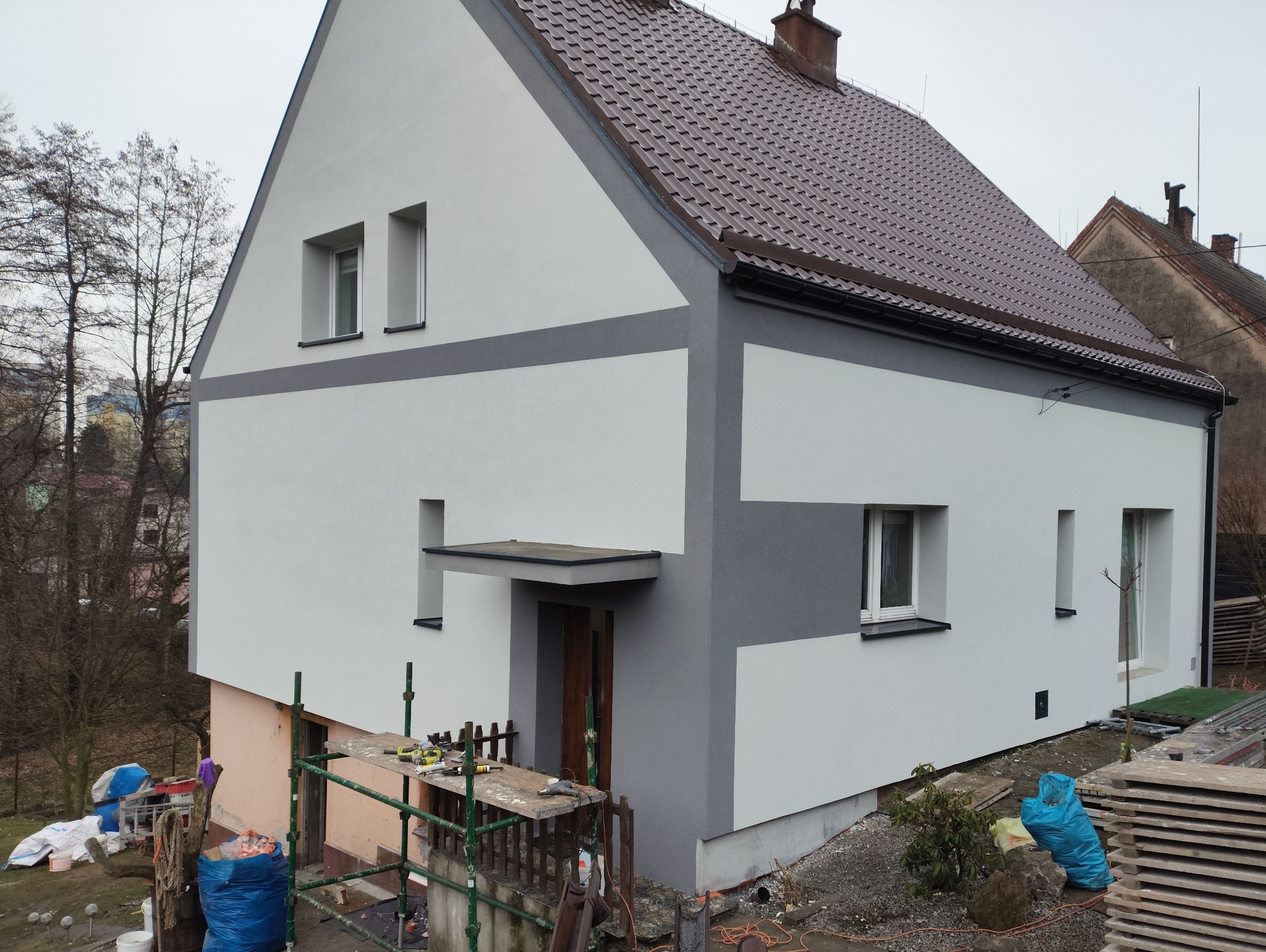 Odnowiona elewacja domu z brązowym dachem, pomalowana na biało i szaro, z widocznym rusztowaniem i narzędziami, sugerującymi trwające prace wykończeniowe.