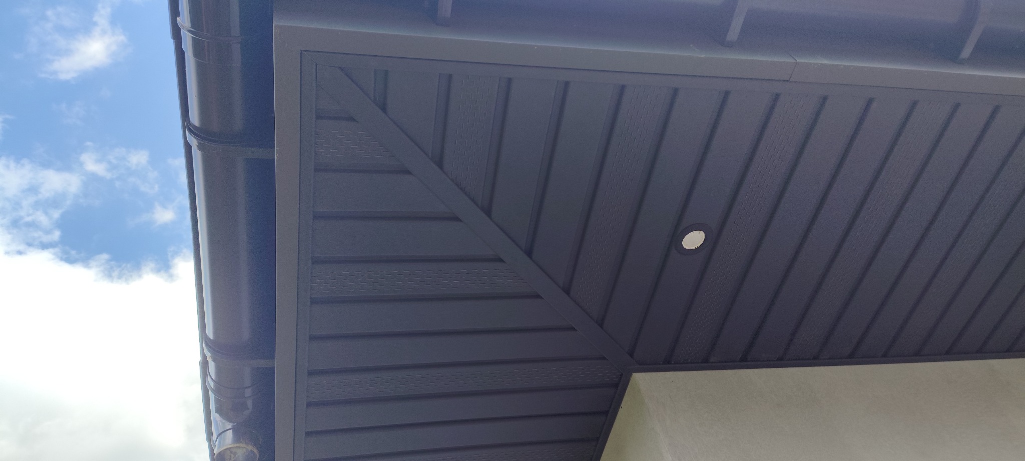 Brunatna podbitka dachowa z oświetleniem punktowym, widok z dołu. Elementy wykończeniowe i rynna na tle błękitnego nieba z chmurami. Estetyczne detale.