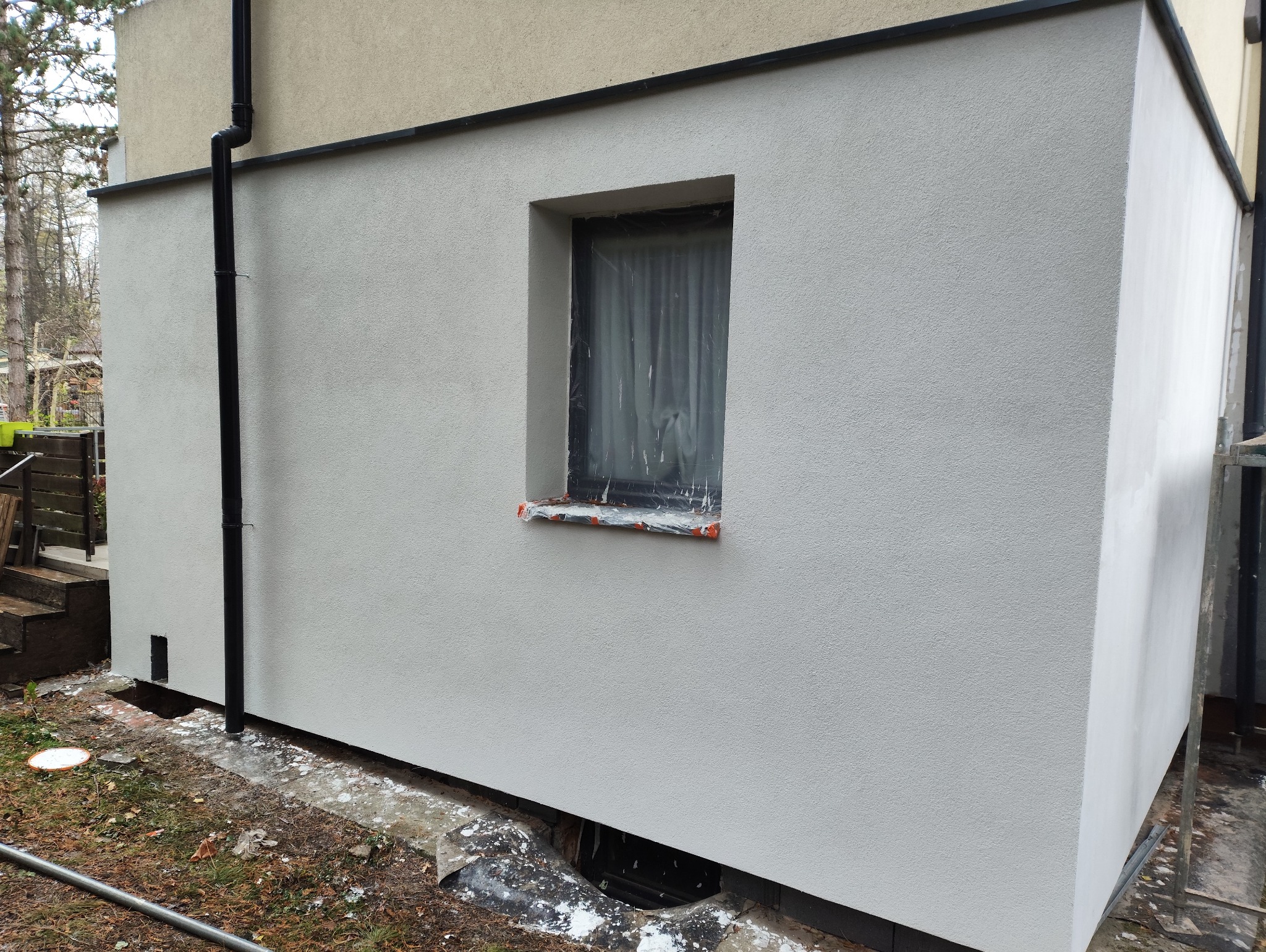 Świeżo otynkowana elewacja budynku z zabezpieczonym oknem folią malarską. Widoczny fragment rynny i ślady prac wykończeniowych na gruncie.
