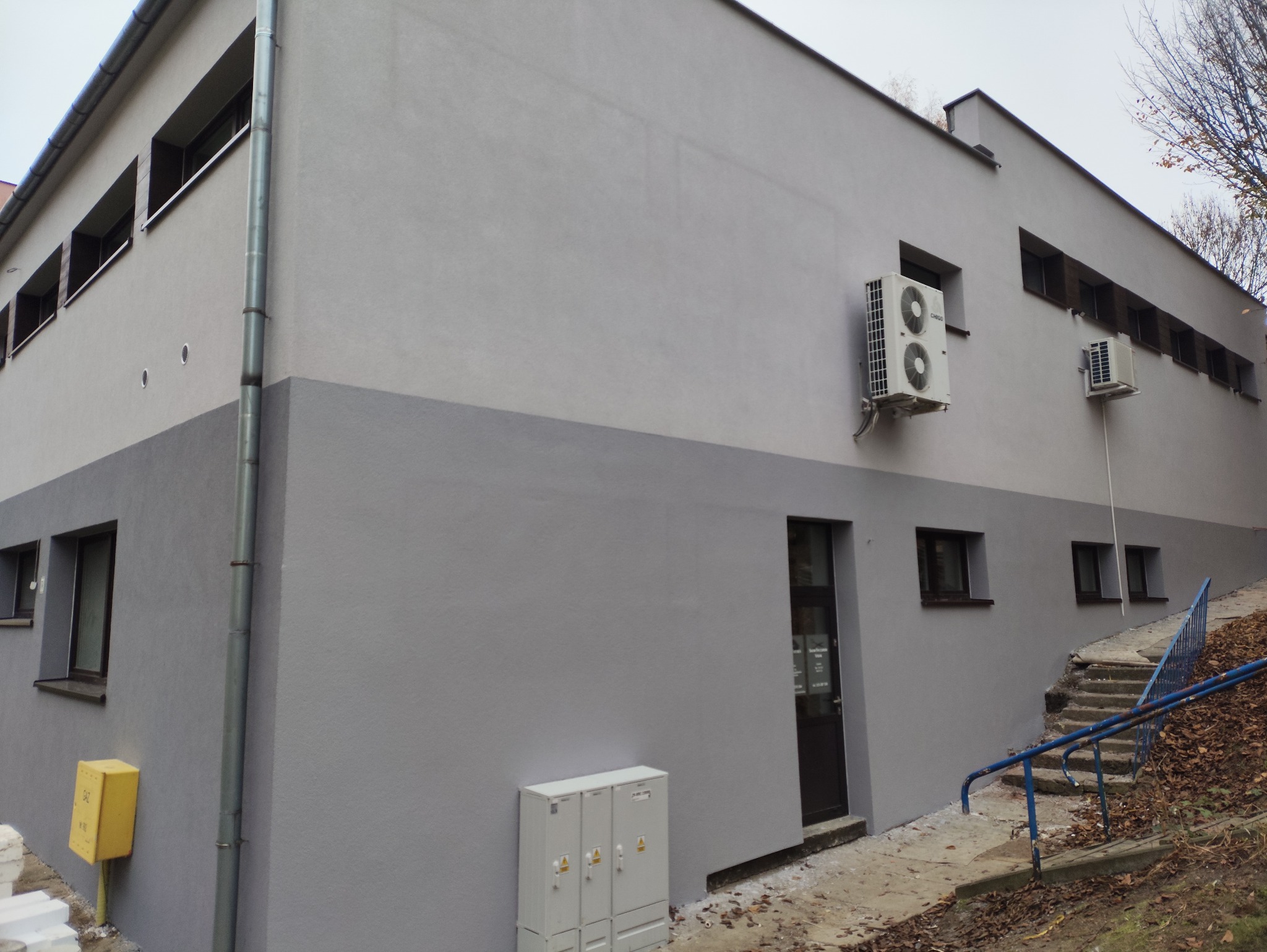 Szara, dwukolorowa elewacja budynku po malowaniu z klimatyzatorami. Widoczne okna, drzwi i schody z niebieską barierką.