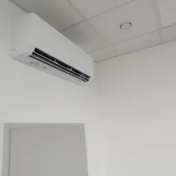 Biały klimatyzator ścienny marki Gree zamontowany w jasnym pomieszczeniu biurowym z kasetonowym sufitem.