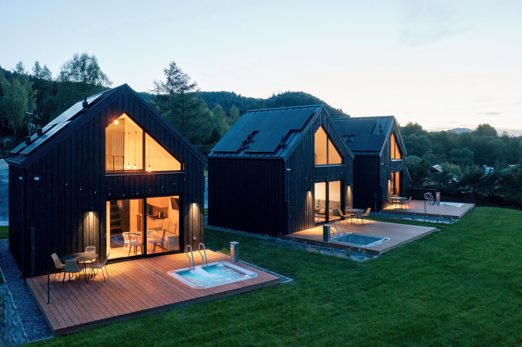 Trzy nowoczesne, czarne domki z drewnianymi tarasami i jacuzzi, oświetlone od wewnątrz, na tle zielonego trawnika i gór o zmierzchu.