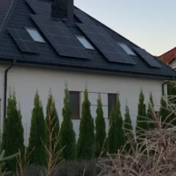 Dom jednorodzinny z panelami słonecznymi na dachu z dachówki, widoczne okna dachowe i komin, ogród z tujami i trawami ozdobnymi na pierwszym planie, sfotografowany o zmierzchu.