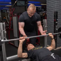 Mężczyzna asekuruje drugiego mężczyznę podczas wyciskania sztangi na ławeczce w siłowni. Widoczny ceglany mur w tle.
