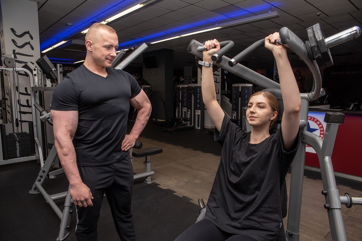Kobieta ćwiczy na maszynie pod okiem instruktora w siłowni w Białymstoku. Widoczny mężczyzna z rozbudowaną muskulaturą obserwujący postępy ćwiczącej.