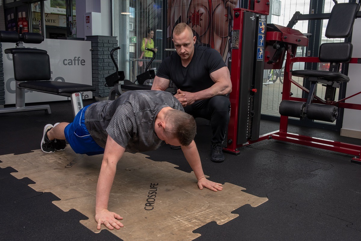Mężczyzna wykonuje pompki na drewnianej macie w siłowni, nadzorowany przez muskularnego mężczyznę w czarnym stroju, widoczny sprzęt do ćwiczeń i elementy wystroju siłowni.