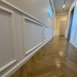 Elegancki korytarz po remoncie: drewniany parkiet ułożony w jodełkę, ściany z białymi panelami dekoracyjnymi i listwami przypodłogowymi. Widok w głąb, okno i oświetlenie.