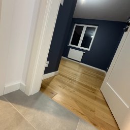 BREMAR Marcel Brejdakowski - Ukończone wnętrze: parkiet w pokoju z granatową ścianą, białe okno i grzejnik. Szare płytki w przejściu, białe drzwi i listwy przypodłogowe.