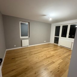BREMAR Marcel Brejdakowski - Jasny, drewniany parkiet w pustym pokoju z szarą ścianą i białymi oknami. Widoczne grzejniki pod oknami. Minimalistyczne wnętrze po remoncie.