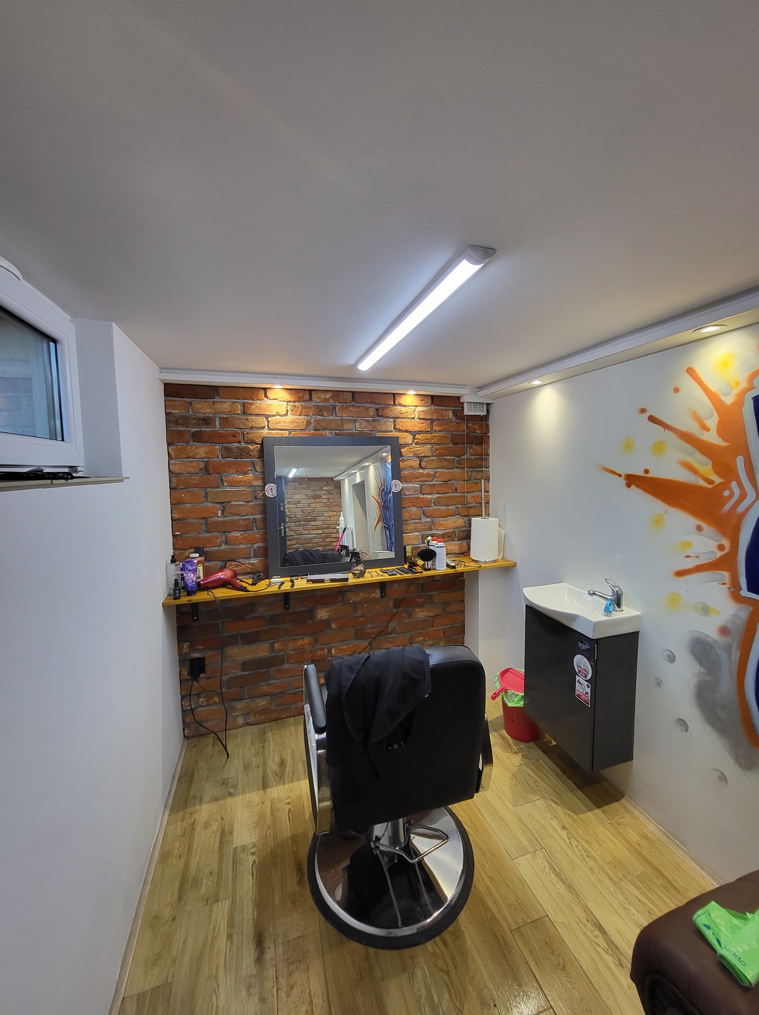 Wnętrze salonu fryzjerskiego z krzesłem, lustrem na tle ceglanej ściany, umywalką i graffiti na ścianie.