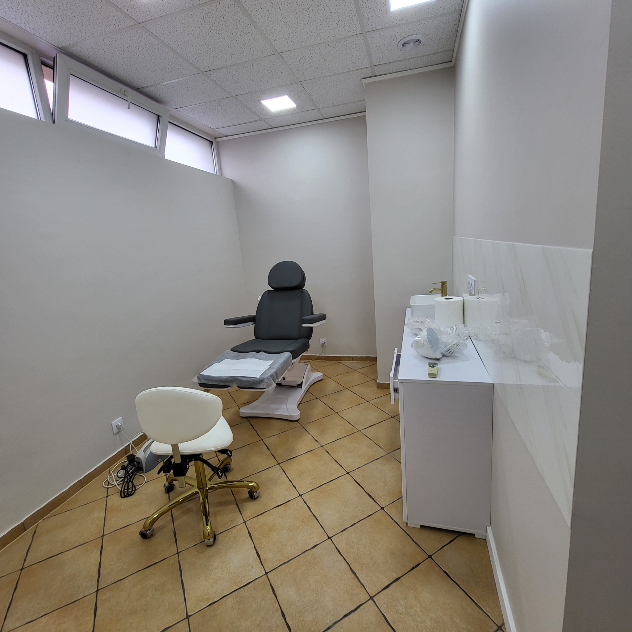 Wnętrze gabinetu kosmetycznego z fotelem zabiegowym, krzesłem na złotych kółkach i umywalką z białym blatem wyłożonym marmurową płytką.