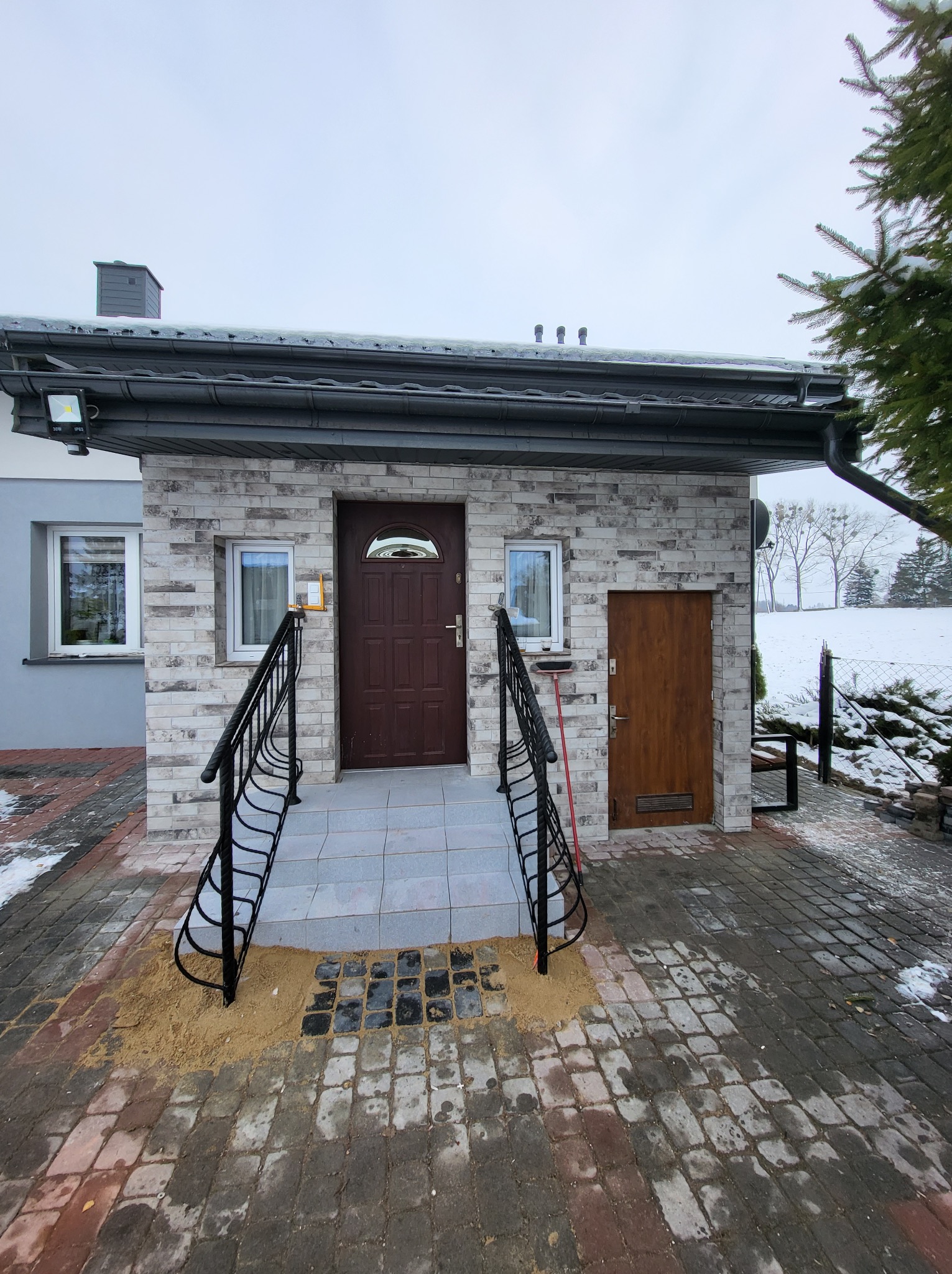 Wejście do domu z drewnianymi drzwiami i schodami wyłożonymi szarą płytką, otoczone elewacją z imitacji kamienia, czarne barierki i kostka brukowa na podjeździe.