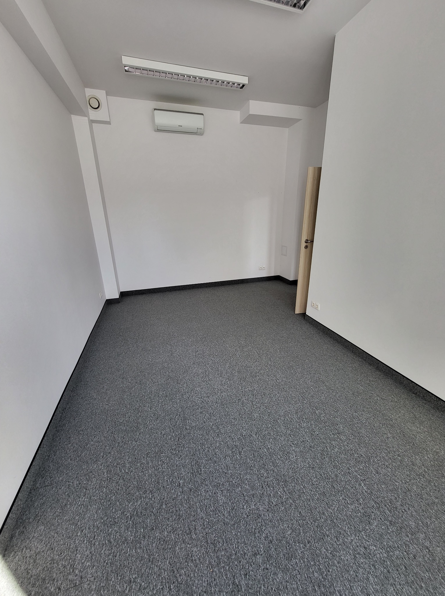 Pusta, jasna przestrzeń biurowa z szarą wykładziną dywanową, klimatyzacją na białej ścianie i drewnianymi drzwiami w tle, oświetlona lampą sufitową.