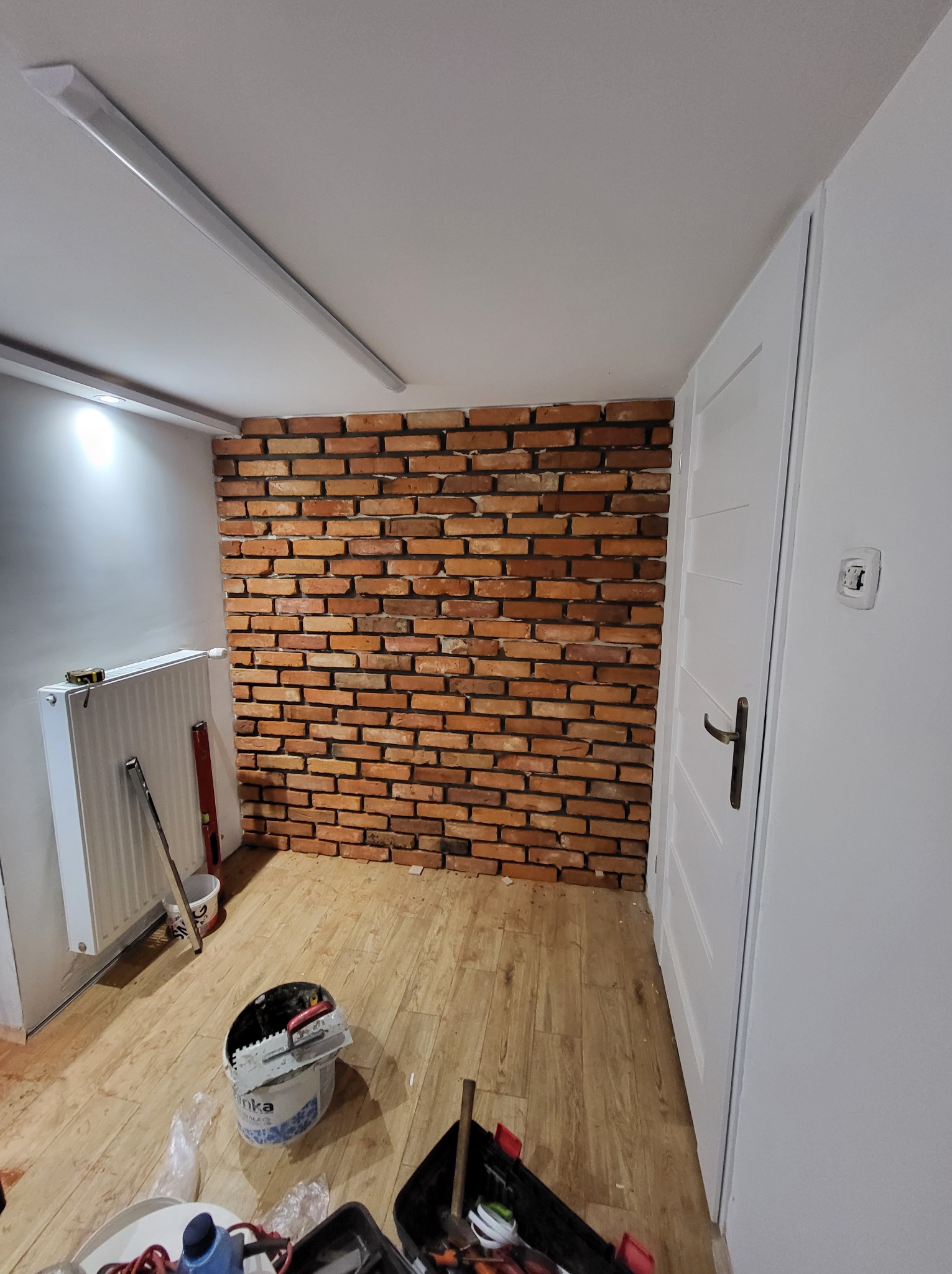 Ściana wykończona cegłą klinkierową w trakcie prac, widoczne narzędzia, wiadro z zaprawą, poziomica, grzejnik i fragment białych drzwi.
