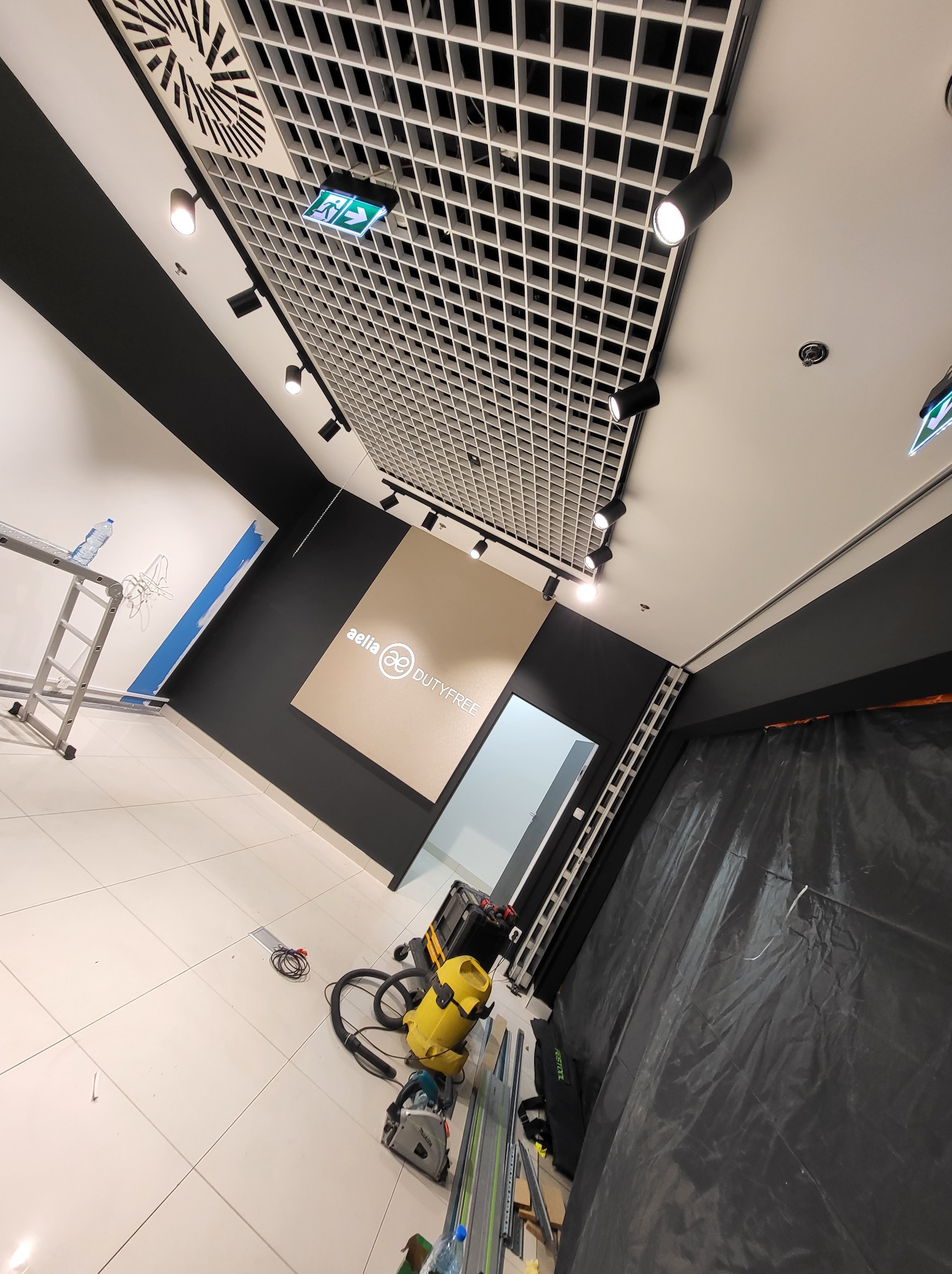 Wnętrze sklepu w trakcie prac wykończeniowych: częściowo pomalowane ściany w kolorach czerni i bieli z logo 'aelia DUTYFREE', zabezpieczona folią strefa, drabina, narzędzia malarskie i odkurzacz...