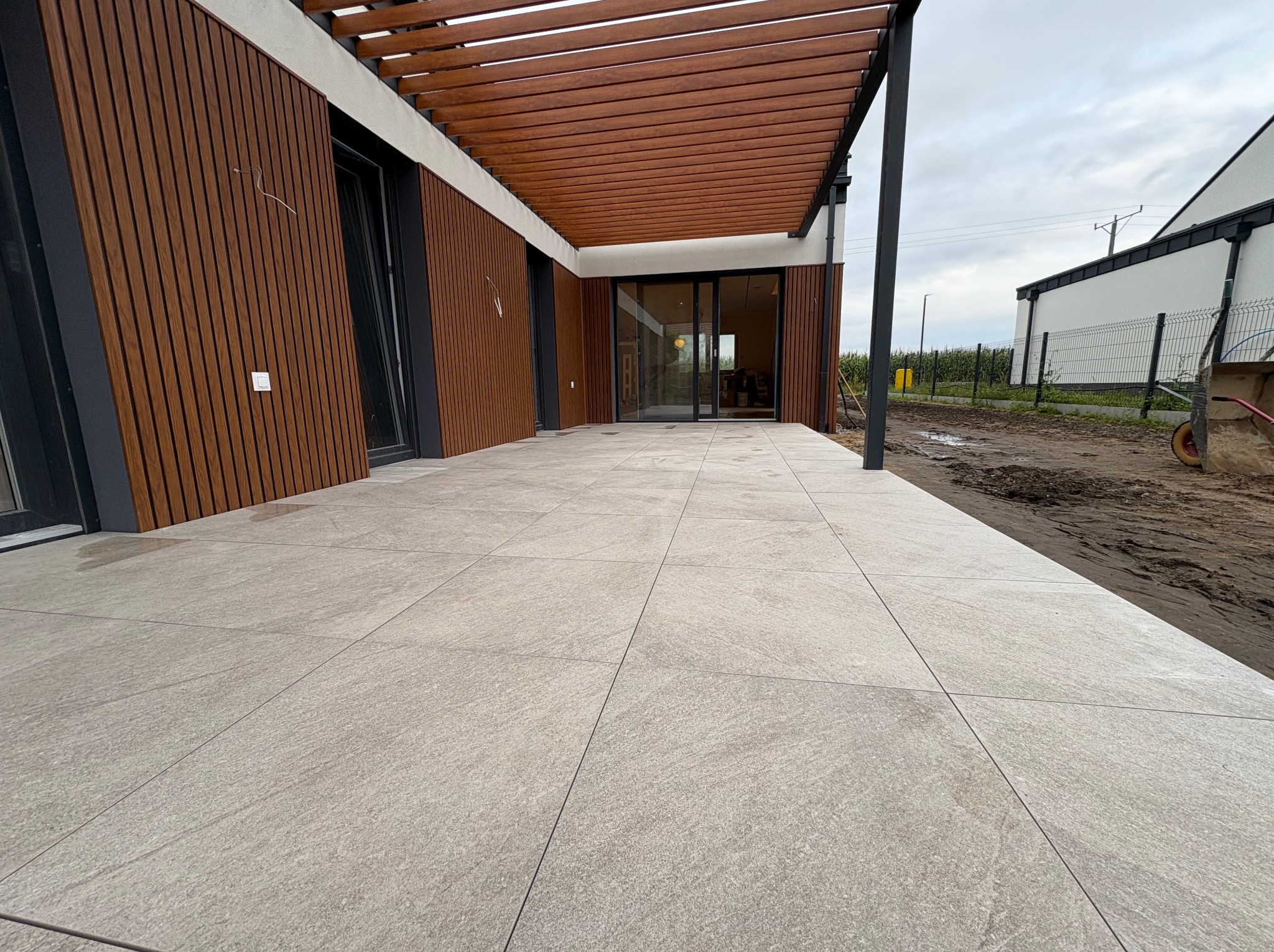 Nowoczesny taras z jasnoszarych płyt na zewnątrz budynku z drewnianą elewacją i pergolą. Widoczny fragment ogrodu i fragment wózka budowlanego.