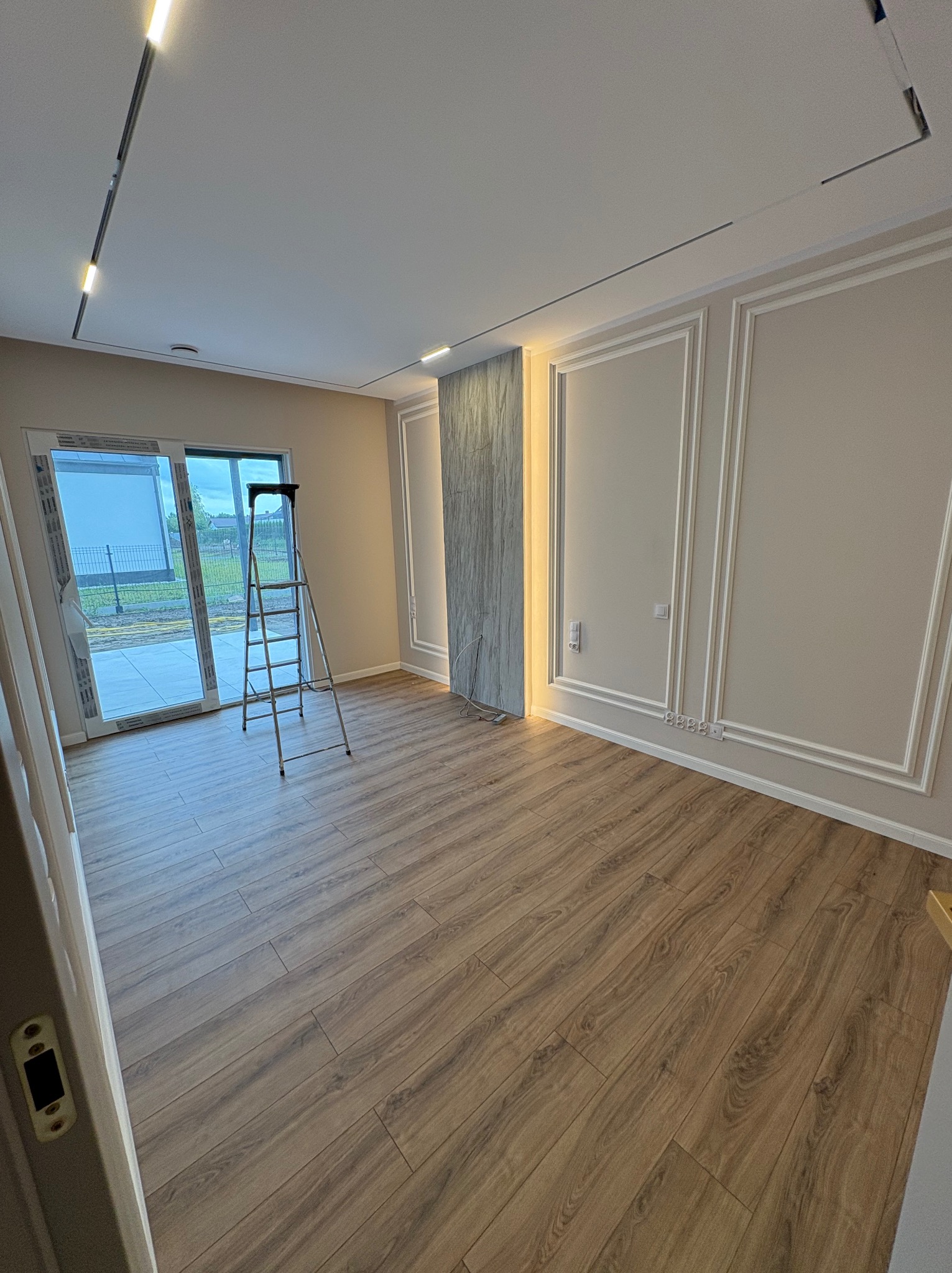 Jasne wnętrze po remoncie z panelami podłogowymi, ozdobnymi listwami na ścianach i nowoczesnym oświetleniem LED. Drabina stoi przy przeszklonym wyjściu na taras.