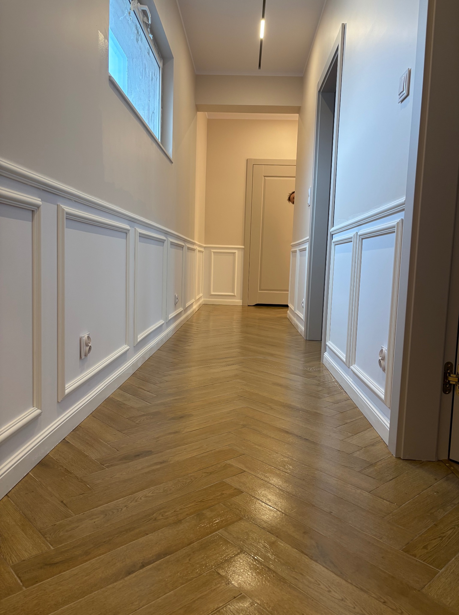 Korytarz po remoncie: parkiet ułożony we wzór jodełki, białe panele ścienne z dekoracyjnymi listwami, jasne ściany i sufit z oświetleniem punktowym.