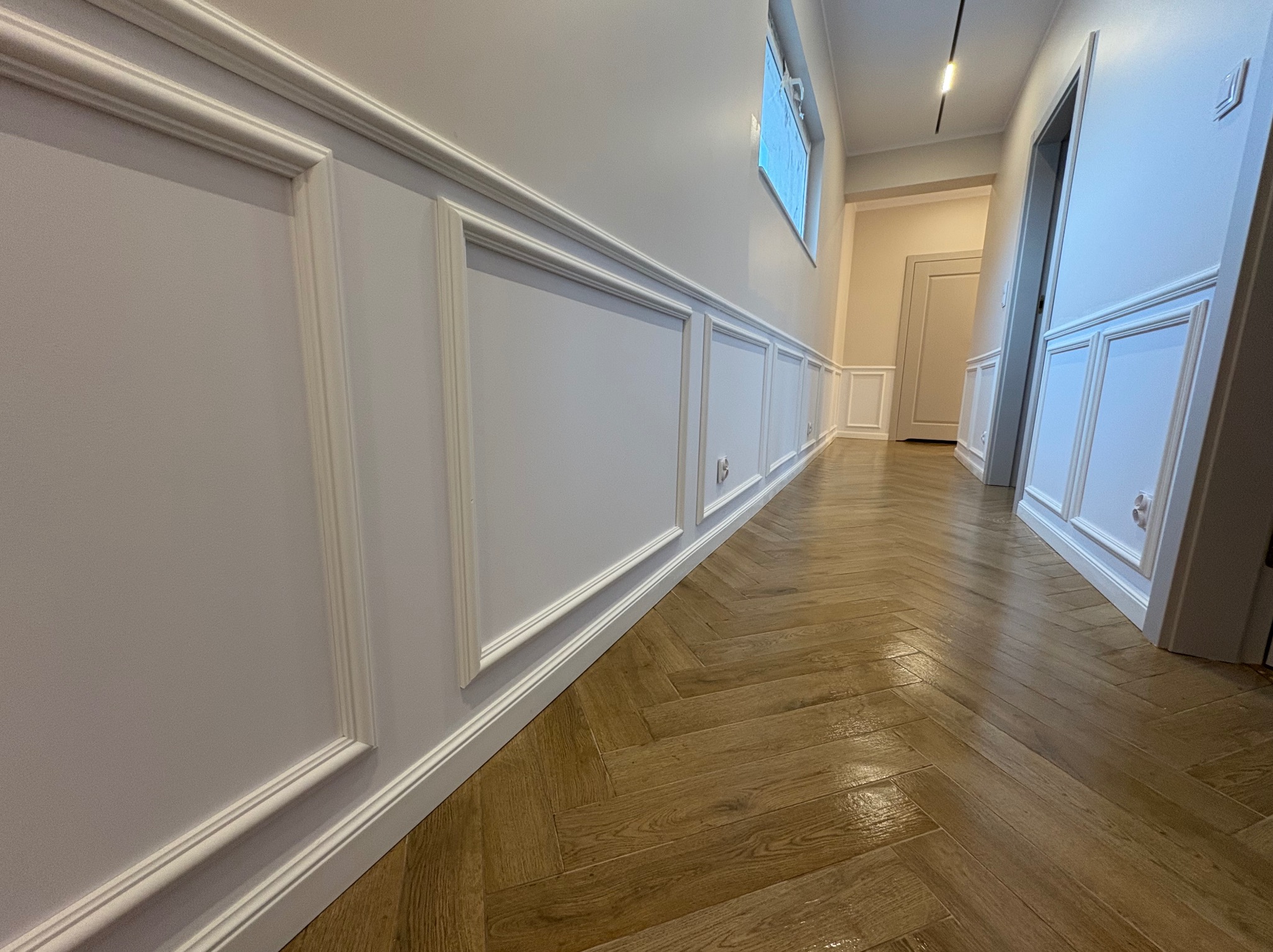 Elegancki korytarz po remoncie: drewniany parkiet ułożony w jodełkę, ściany z białymi panelami dekoracyjnymi i listwami przypodłogowymi. Widok w głąb, okno i oświetlenie.