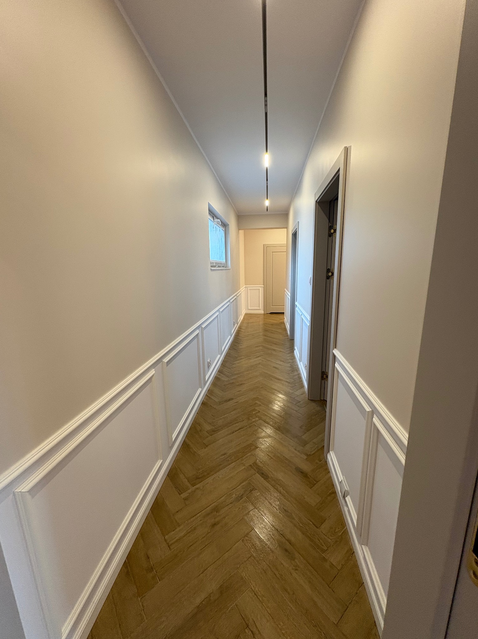 Wykończony korytarz z parkietem w jodełkę, ścianami z panelami dekoracyjnymi i nowoczesnym oświetleniem szynowym. Widok w perspektywie, podkreślający długość i elegancję wnętrza.