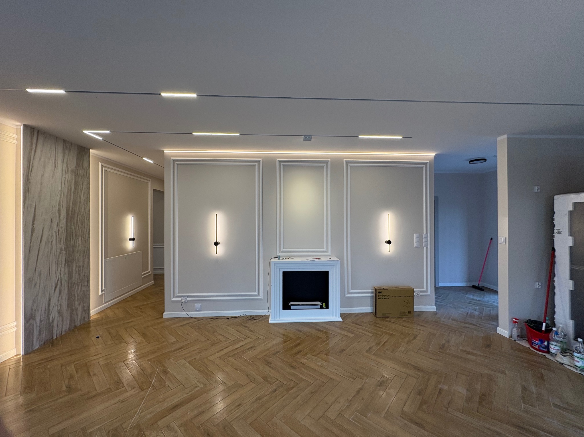 Eleganckie wnętrze po remoncie: parkiet w jodełkę, ściany w sztukaterii z dyskretnym oświetleniem, imitacja kominka. Widoczne ślady po remoncie, kartony i farby.