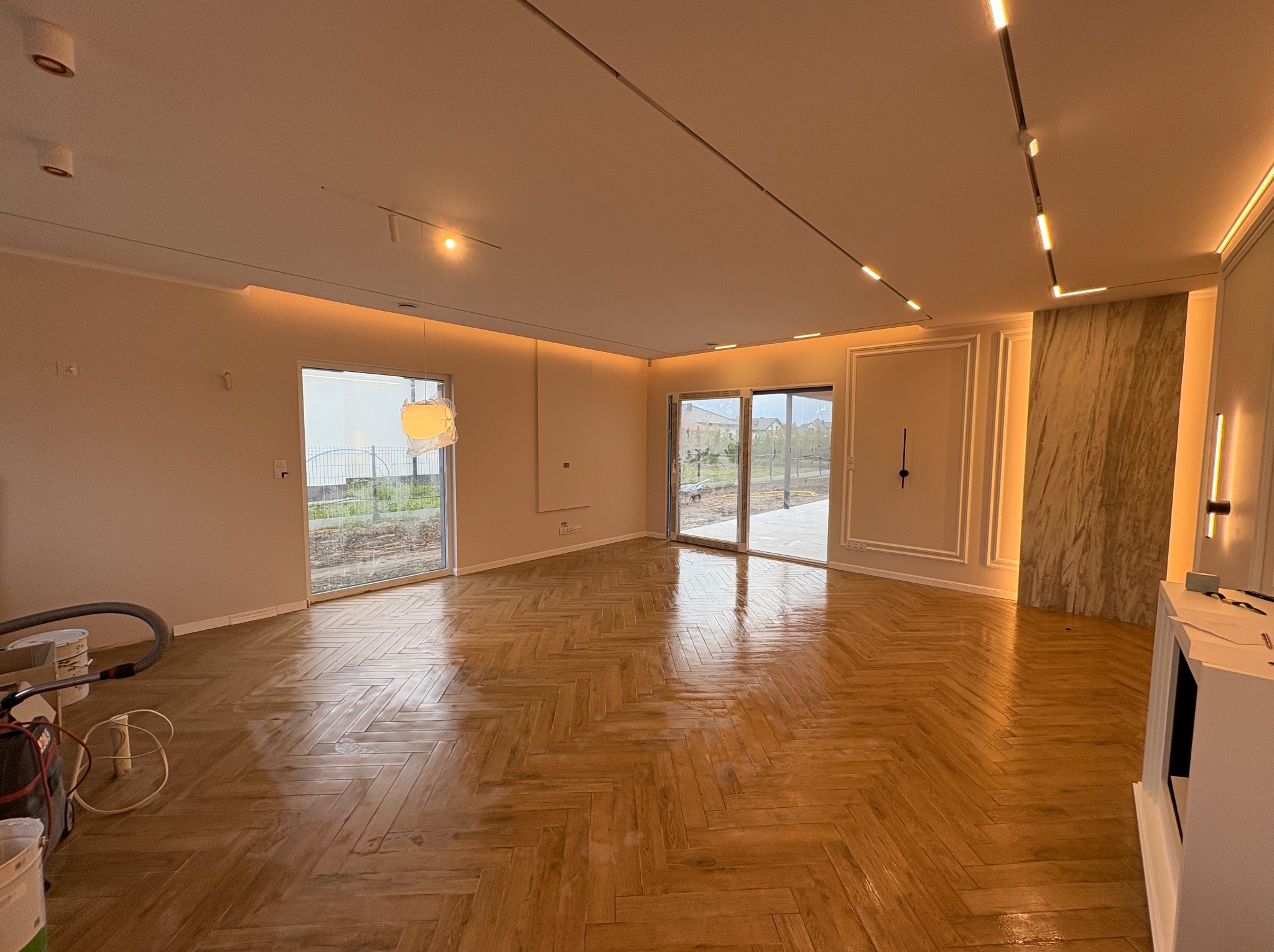 Salon po remoncie: parkiet w jodełkę, jasne ściany, przesuwne drzwi balkonowe, oświetlenie ledowe. Widoczne narzędzia pracy.