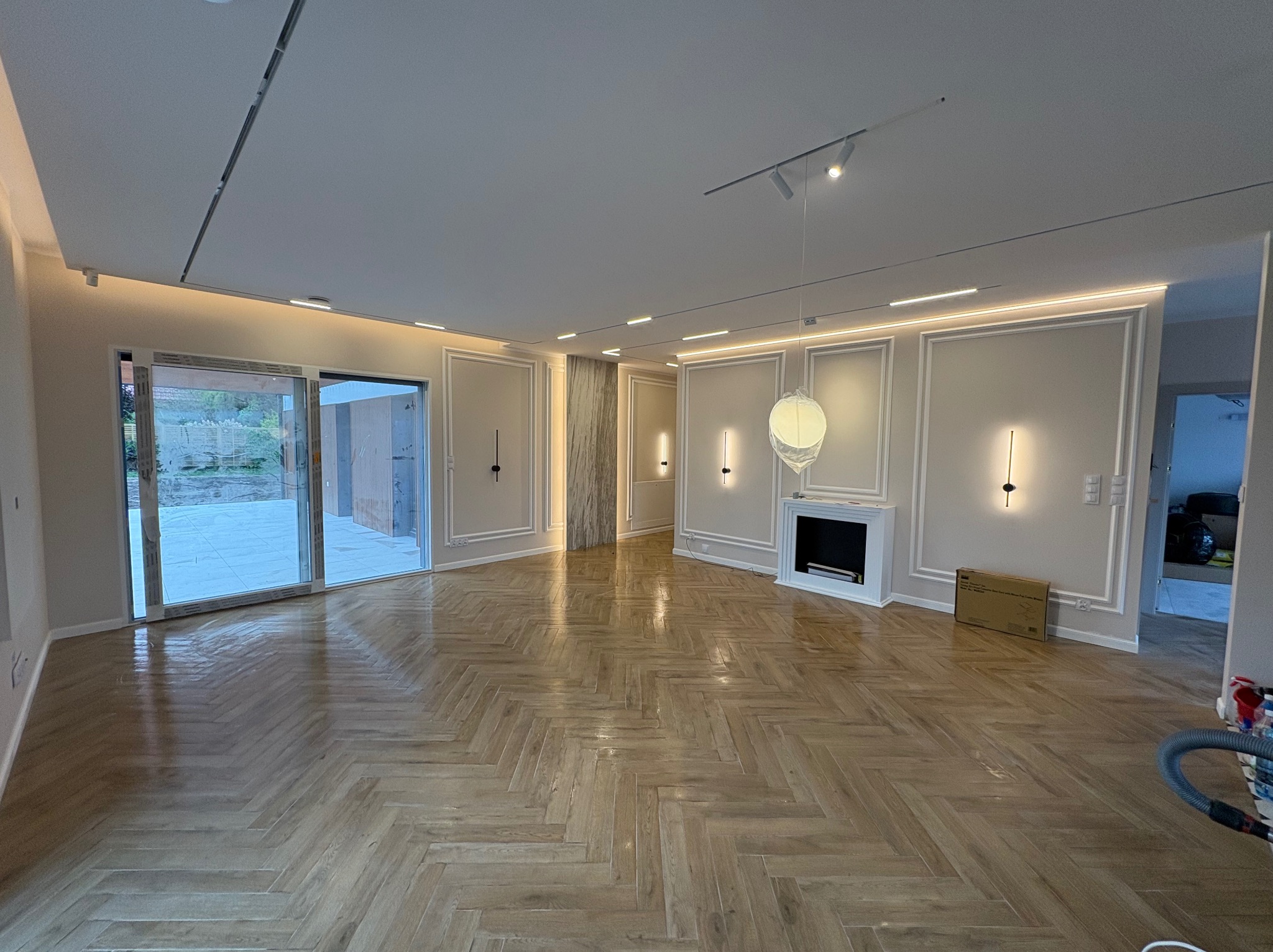 Jasne, przestronne wnętrze po remoncie z parkietem w jodełkę, listwami ściennymi i nowoczesnym oświetleniem. Widoczne przesuwne drzwi balkonowe i elementy dekoracyjne.