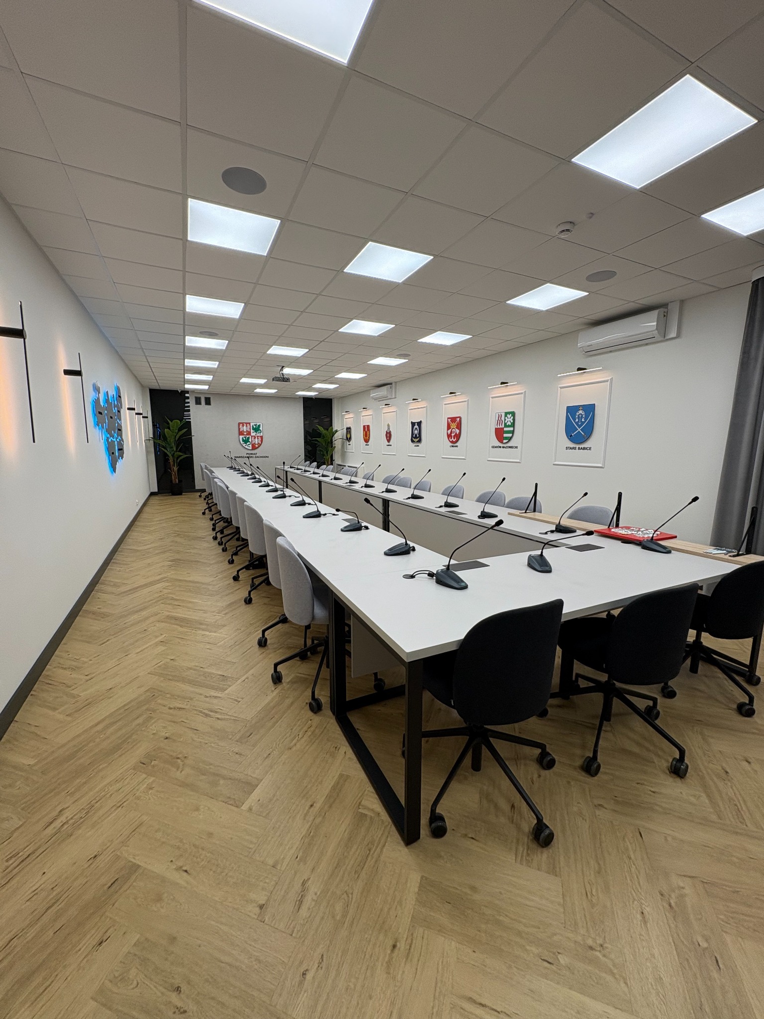 Nowoczesna sala konferencyjna z długim białym stołem, szarymi krzesłami i parkietem w jodełkę. Na ścianach herby i podświetlana mapa. Klimatyzacja i oświetlenie sufitowe.