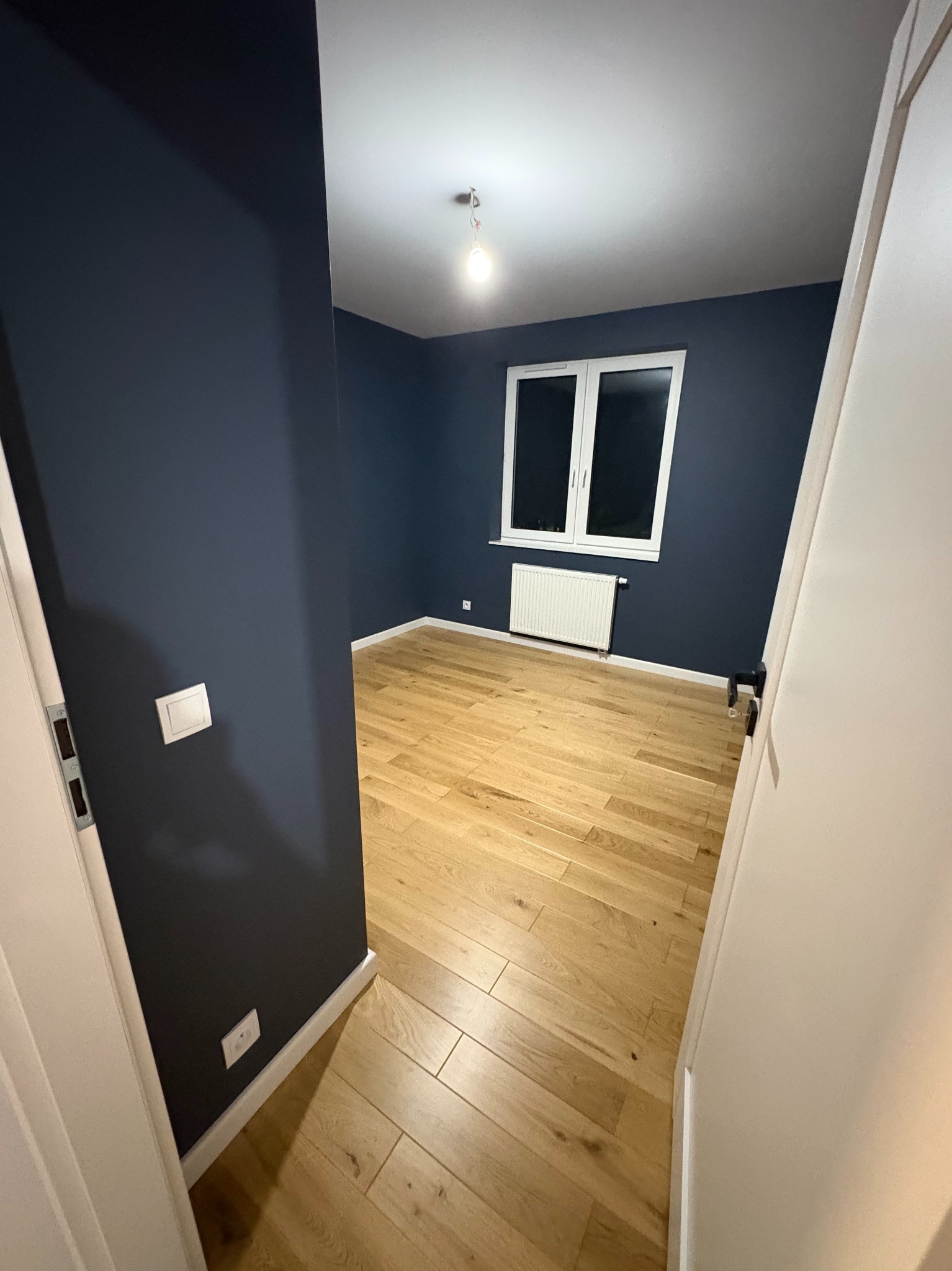 Pokój po remoncie: drewniana podłoga, ściany w odcieniach granatu, białe okno i grzejnik. Widok zza otwartych, białych drzwi.