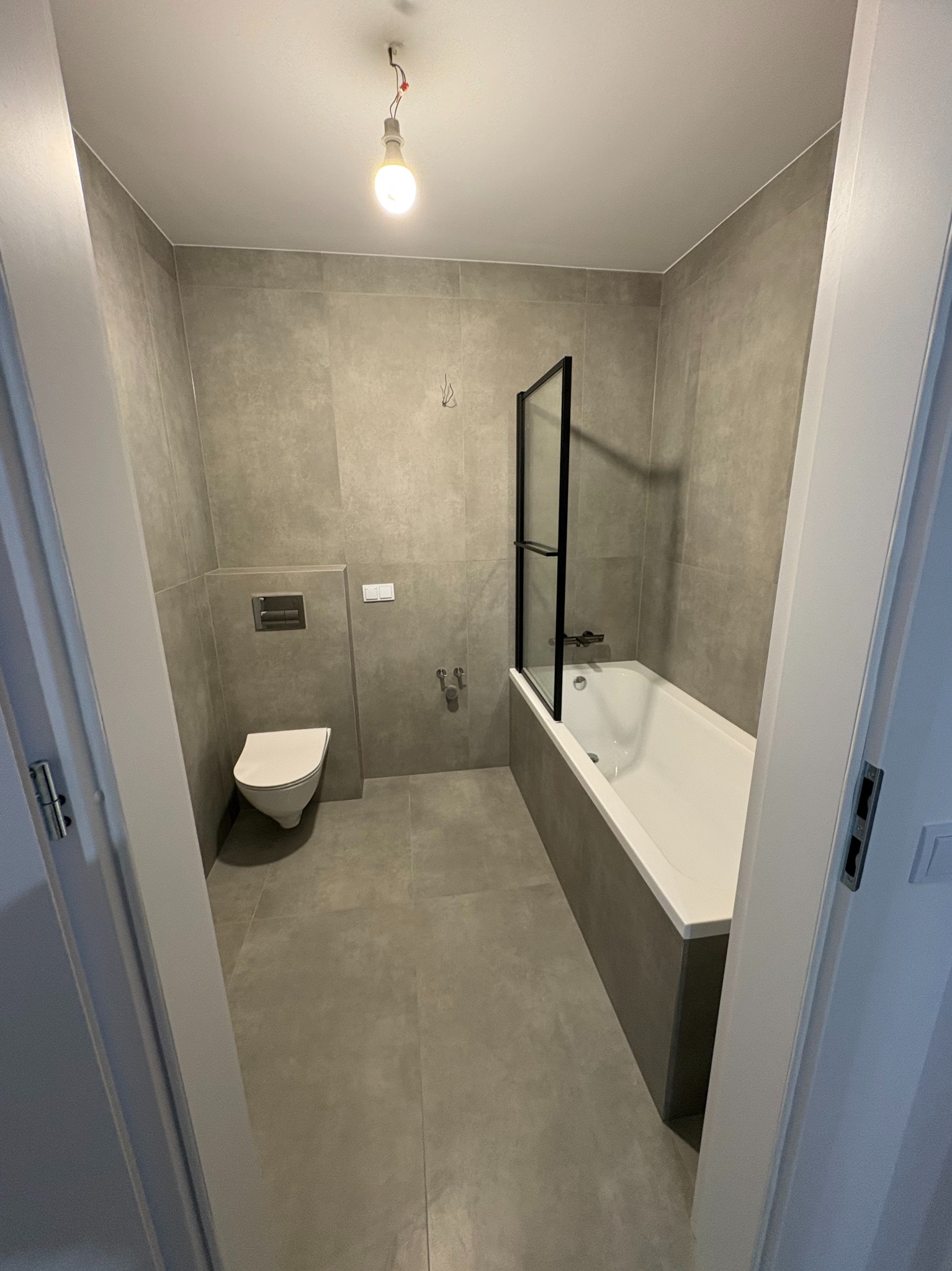 Minimalistyczna łazienka w szarościach: płytki imitujące beton, biała wanna z czarnym parawanem, podwieszana toaleta. Surowy, nowoczesny design.