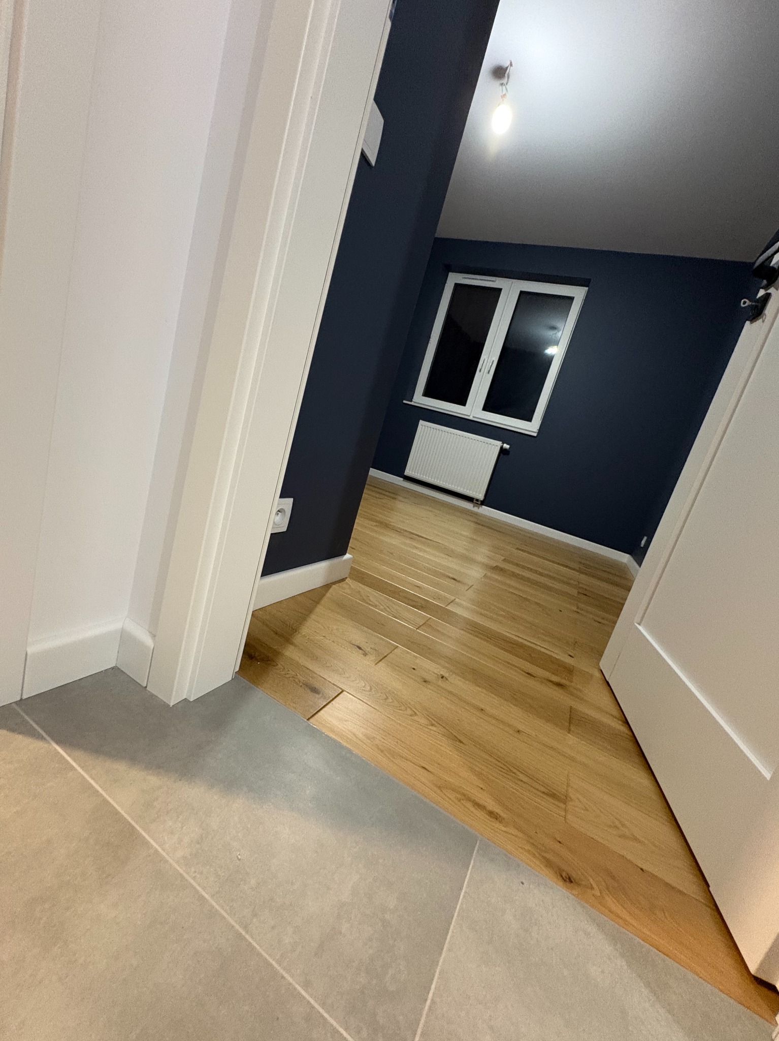 Ukończone wnętrze: parkiet w pokoju z granatową ścianą, białe okno i grzejnik. Szare płytki w przejściu, białe drzwi i listwy przypodłogowe.