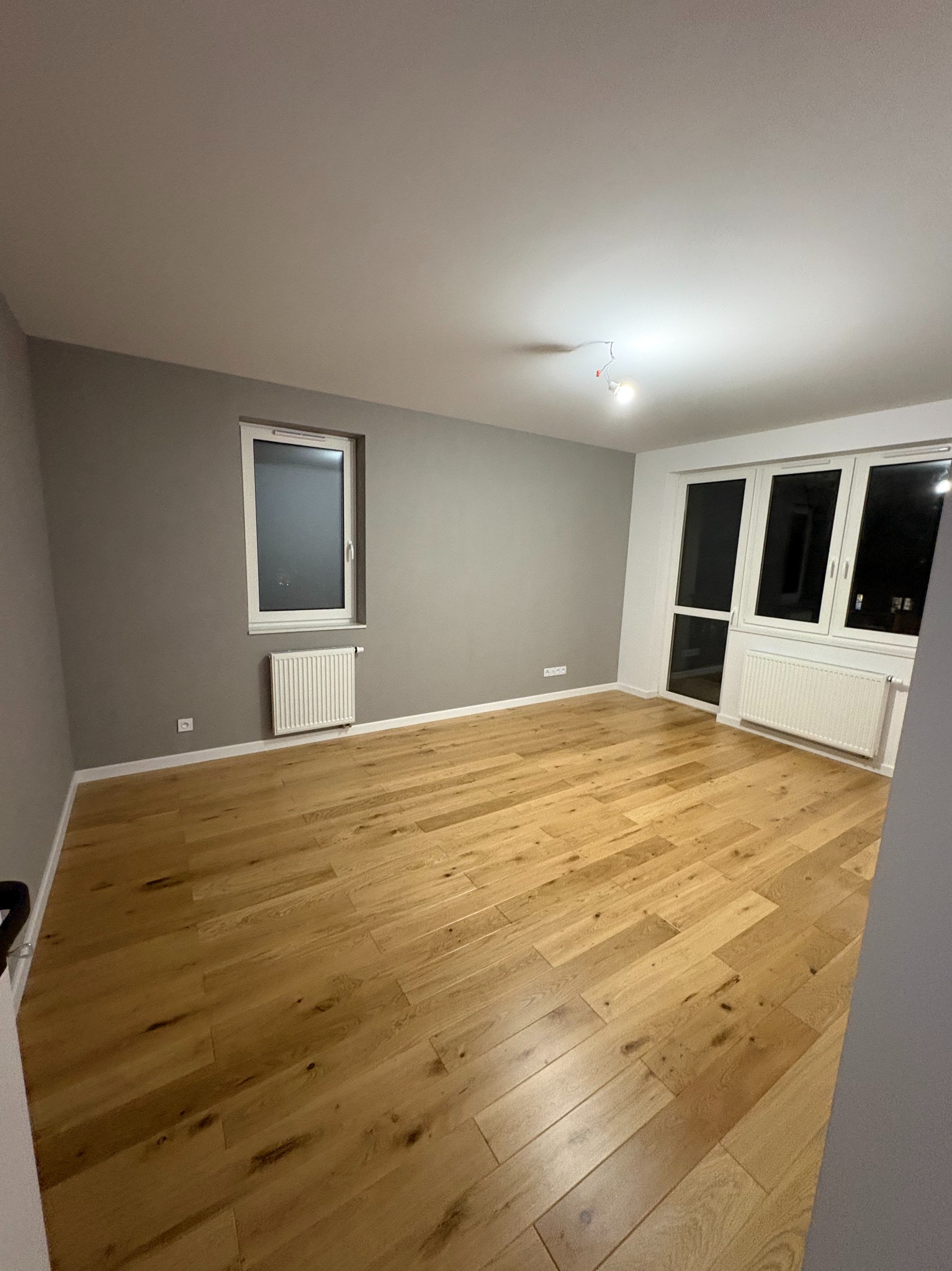 Jasny, drewniany parkiet w pustym pokoju z szarą ścianą i białymi oknami. Widoczne grzejniki pod oknami. Minimalistyczne wnętrze po remoncie.