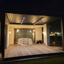 Nowoczesna, oświetlona pergola z drewnianą podłogą na tle nocnego krajobrazu miejskiego, stół i krzesła przykryte folią ochronną.