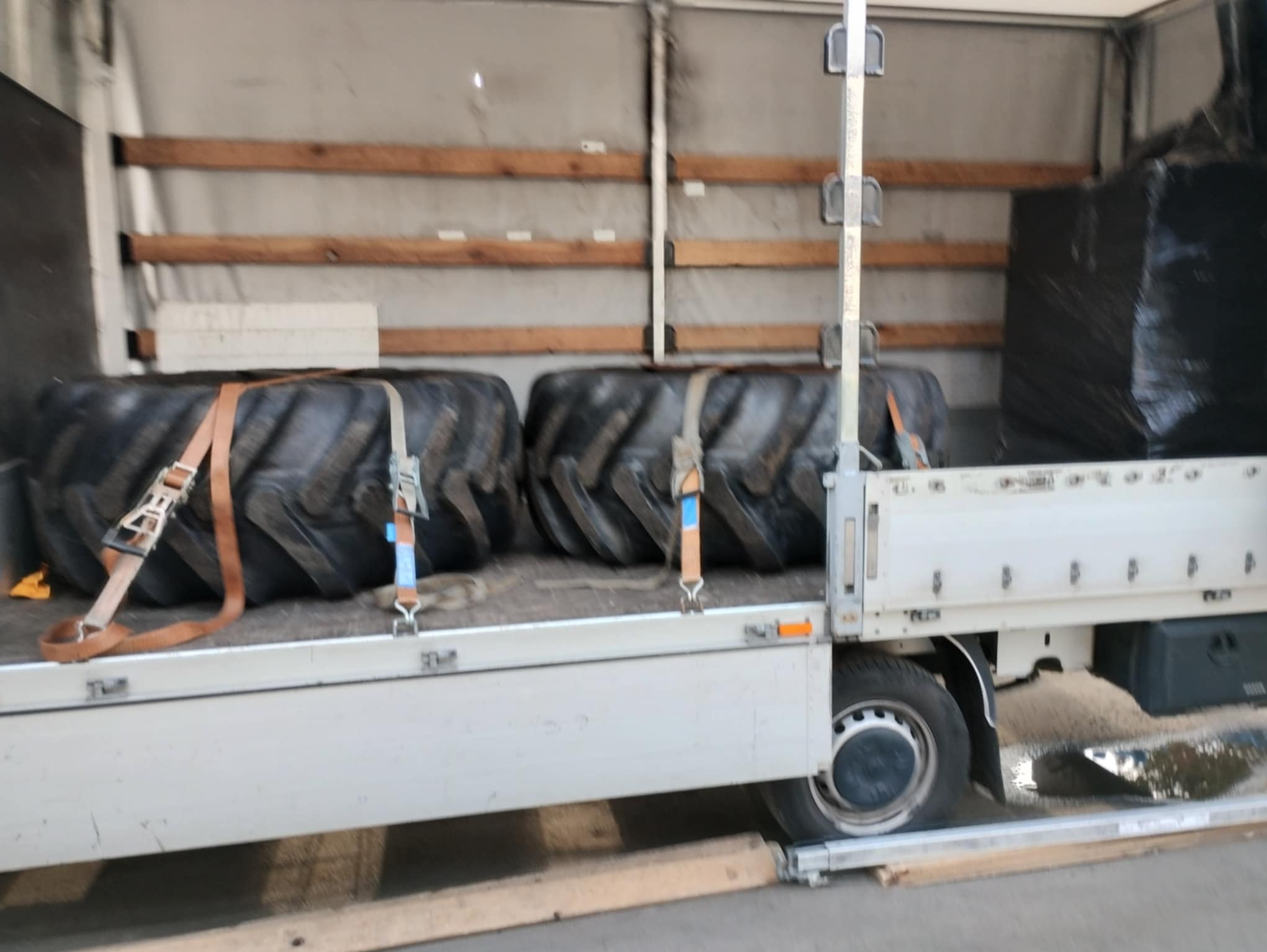 Wnętrze busa z dwoma dużymi oponami zabezpieczonymi pasami transportowymi. W tle widoczne paczki owinięte folią stretch.