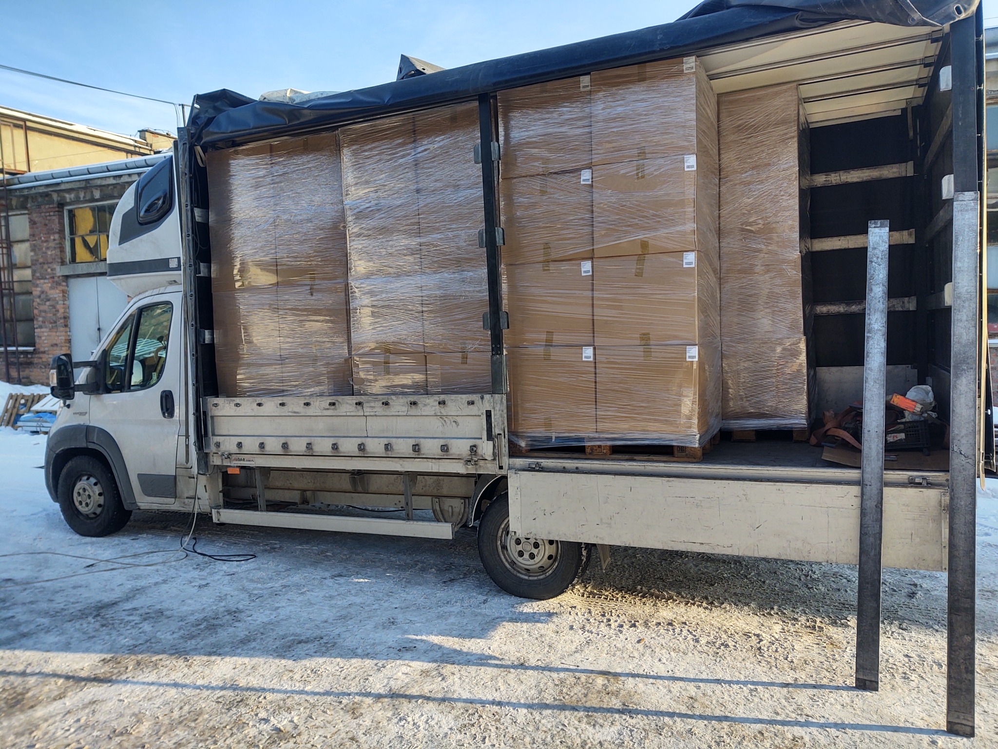 Ciężarówka dostawcza załadowana kartonami owiniętymi folią, gotowa do transportu. Widoczne palety i zabezpieczenia ładunku. Zimowy krajobraz w tle.