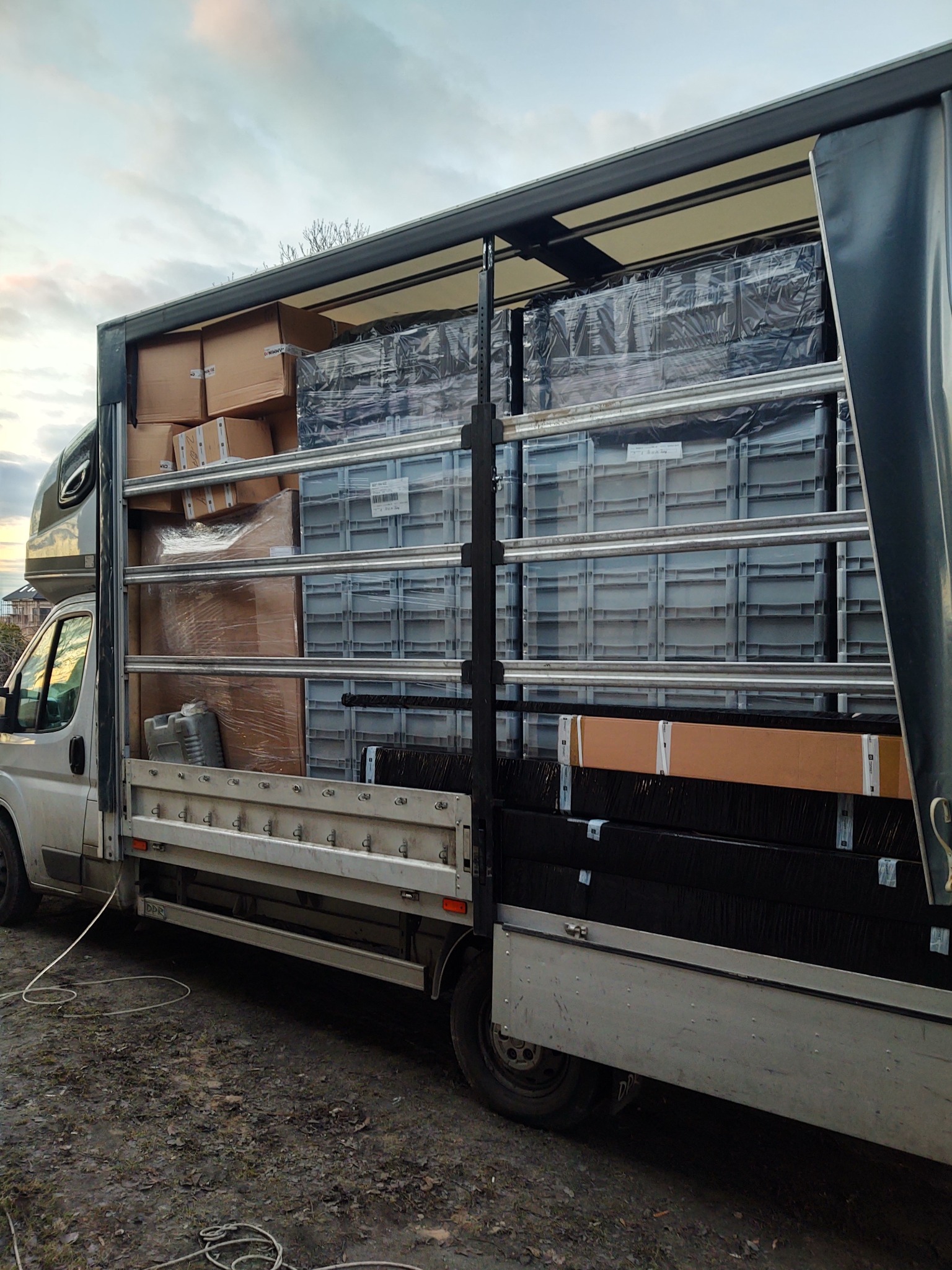 Bus z otwartą plandeką, wypełniony kartonami i plastikowymi pojemnikami, zabezpieczonymi folią. Widok z boku na załadowany pojazd na zewnątrz.