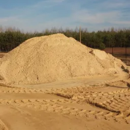 Duża pryzma piasku z wyraźnymi śladami opon pojazdu budowlanego, tło stanowią drzewa i błękitne niebo.