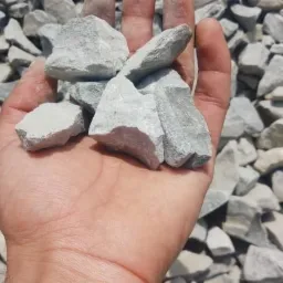 Dłoń trzymająca jasnoszare, nieregularne bryły kruszywa na tle większej sterty podobnych kamieni.