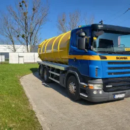 Niebiesko-żółty samochód ciężarowy Scania R420 z cysterną na chodniku z kostki, w tle budynek i drzewa bez liści pod błękitnym niebem.