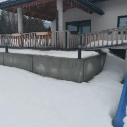 Betonowy fundament domu jednorodzinnego z drewnianym tarasem i zjeżdżalnią dla dzieci, pokryty śniegiem w zimowej scenerii.