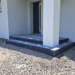 Wykończone dwustopniowe schody wejściowe do domu z szarej kostki brukowej, z kolumną podtrzymującą zadaszenie, otoczone żwirem.