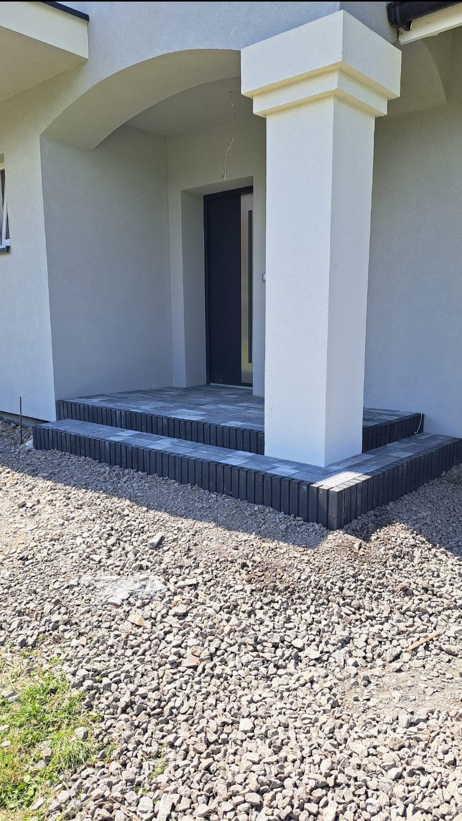 Wykończone dwustopniowe schody wejściowe do domu z szarej kostki brukowej, z kolumną podtrzymującą zadaszenie, otoczone żwirem.