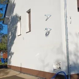 Świeżo pomalowana, biała elewacja domu jednorodzinnego z brązowym cokołem, widoczne uchwyty na kable i rynna, na ziemi leżą narzędzia i wąż.